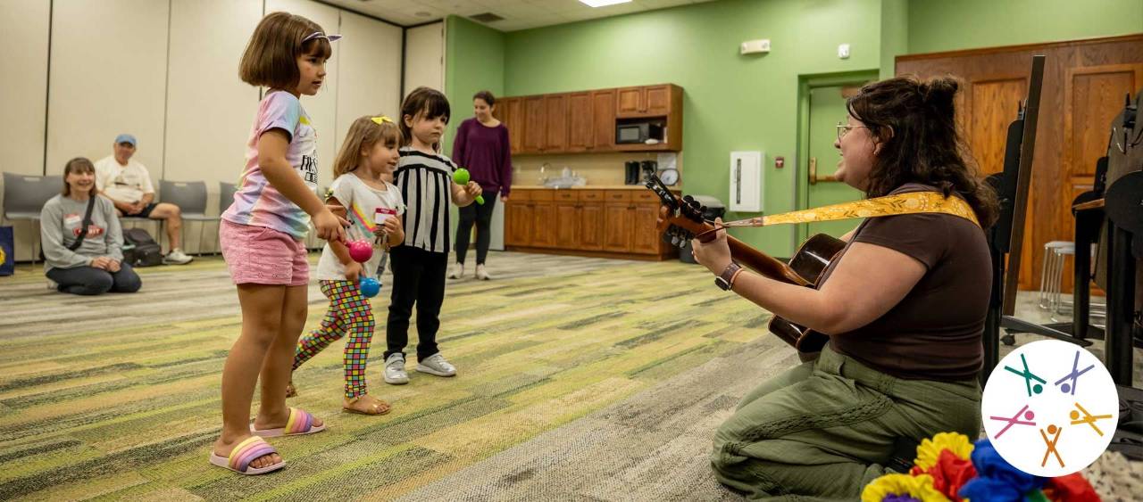 children stand listening to a woman sitting on the ground playing guitar
